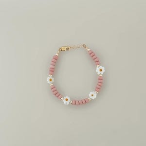 May include: A pink beaded bracelet with white daisy flower accents and gold-colored metal details. The bracelet is made of small, round beads and features a clasp closure. The daisies have yellow centers.