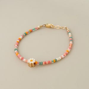 May include: A colorful beaded bracelet with a gold clasp and a daisy charm. The bracelet features small, round beads in shades of pink, orange, green, and white. The daisy charm has a white flower with a yellow center and a gold base.