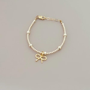 May include: Gold-toned bracelet featuring small, round beads and a delicate bow charm. The bow charm has a small, clear gemstone. The bracelet is secured with a lobster clasp and an adjustable chain.