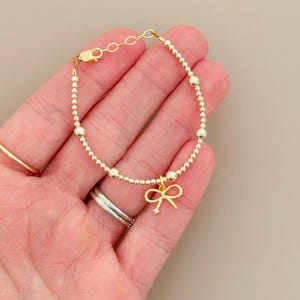 May include: A gold-colored bracelet with a delicate bow charm. The bracelet features small, round beads and a lobster clasp closure. The bow charm has a small, clear gemstone in the center. The bracelet is shown on a hand.