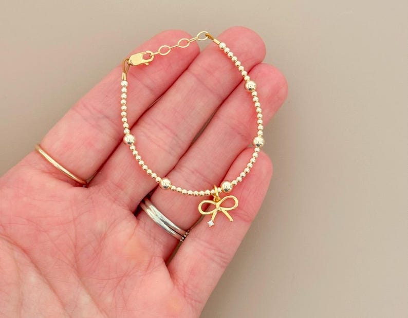 May include: A gold-colored bracelet with a delicate bow charm. The bracelet features small, round beads and a lobster clasp closure. The bow charm has a small, clear gemstone in the center. The bracelet is shown on a hand.