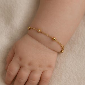 May include: A baby's hand adorned with a fine gold bracelet. The bracelet is composed of small, spherical beads linked by a delicate chain. The skin is pale, and the backdrop is a soft, neutral tone.