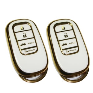 May include: Two white car key fobs with gold trim. Each fob has four buttons labeled with icons for lock, unlock, trunk, and panic. The buttons also have the word "HOLD" printed on them.
