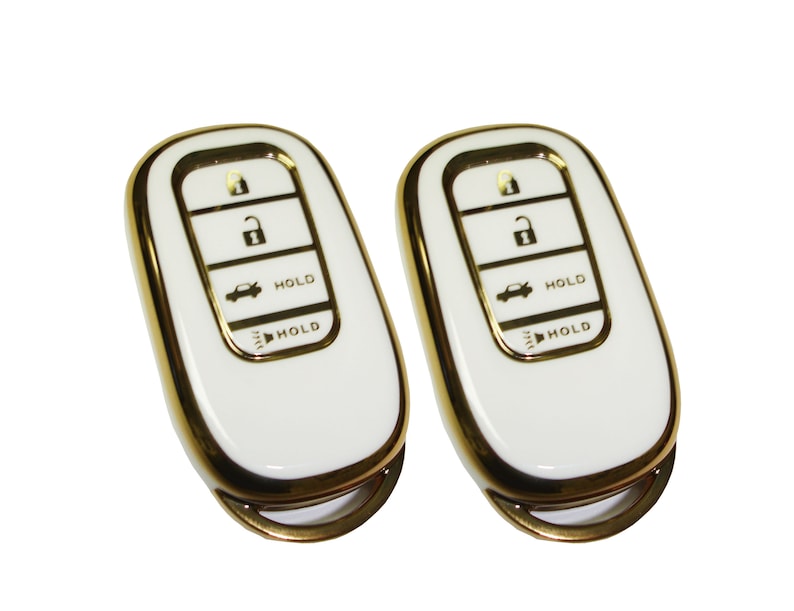May include: Two white car key fobs with gold trim. Each fob has four buttons labeled with icons for lock, unlock, trunk, and panic. The buttons also have the word "HOLD" printed on them.