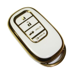 May include: White car key fob with a gold trim. The key fob has four buttons: unlock, lock, trunk release, and panic. The buttons are labeled with the words "HOLD".