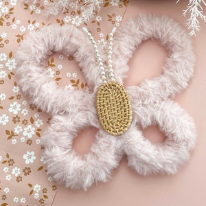 May include: A decorative butterfly made from soft, light pink material. The circular wings are joined by a woven, oval body. White pearl beads form the antennae. The butterfly sits on a pink floral patterned background.