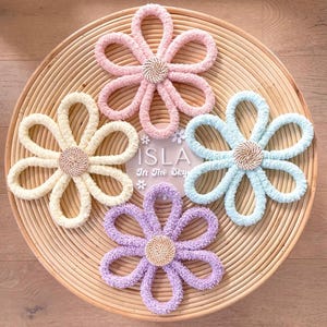 Può includere: Tre decorazioni murali a forma di fiore all'uncinetto in colori pastello: rosa chiaro, azzurro e lavanda. Ogni fiore ha un centro intrecciato ed è esposto su uno sfondo in vimini intrecciato. Il testo "Isla In The Sky" è visibile su un cerchio bianco al centro dell'immagine.