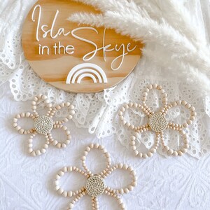 Puede incluir: Un letrero de madera con el texto "Isla Skye in the" y un recorte de arcoíris blanco. Tres decoraciones de flores de cuentas de madera con un centro tejido.