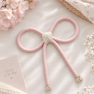 May include: A decorative bow crafted from light pink yarn wrapped around a flexible core. The centre features a cluster of small, clear, shimmering beads. Two long tails extend downward, each finished with a small beige tassel and a gold-toned charm.