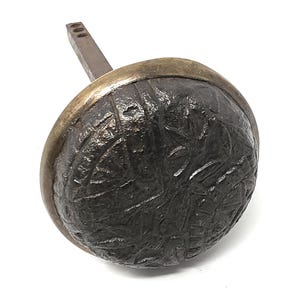 May include: A round door knob featuring a dark, textured, weathered iron face with an embossed geometric pattern and a smooth, polished brass-toned metal rim. A square-shaped metal spindle extends from the back of the knob for installation.