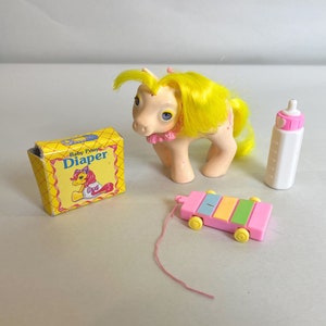 Puede incluir: Un juguete de pony bebé amarillo y rosa con un lazo rosa, una caja de pañales amarilla con el texto "Baby Pony Diaper", un xilófono de juguete rosa y blanco, y una botella blanca con una tapa rosa.