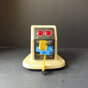 May include: A vintage desk vacuum cleaner toy in a cream color with a black base. It has two red circular lights and the word "DIBOT" printed on the front. A small yellow and blue brush is attached to the front.