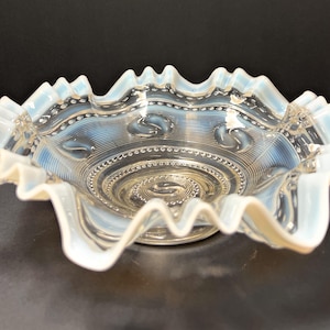 May include: A clear, opalescent glass bowl with a ruffled edge. The bowl features a spiral design in the center and a beaded pattern around the rim. The edges have a wavy, scalloped design.