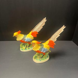 May include: Two ceramic pheasant figurines. The birds have yellow, orange, blue, and green plumage. Their long tail feathers are beige with a dotted pattern. Each bird is perched on a green leaf-shaped base.
