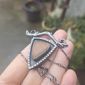 May include: A silver-toned pendant necklace featuring a triangular design with a light brown stone center. The pendant has a textured border and is suspended from a delicate chain. The design includes a branch-like element at the top.