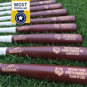 May include: A collection of personalized baseball bats with white handles and brown barrels, engraved with names, numbers, and "Class of 2020." The bats are arranged on green grass, with a "Most Popular" badge.