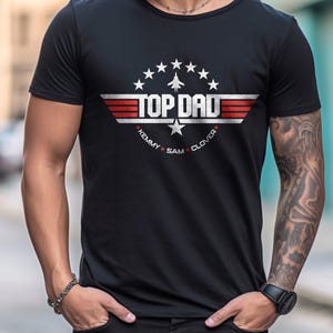 May include: Black t-shirt with a white graphic of a fighter jet and the text "TOP DAD" with the names "KEMMY SAM CLOVER" underneath.