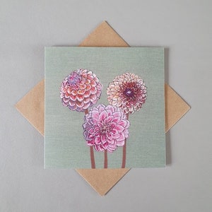 May include: A green card with three pink and white dahlia flowers illustrated on it. The card is surrounded by a brown envelope.