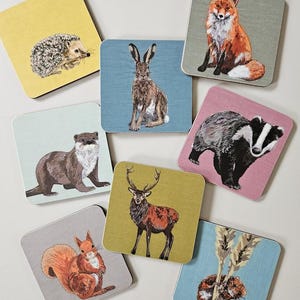 May include: A set of square coasters with illustrations of British wildlife. The coasters depict a hedgehog, fox, hare, otter, badger, squirrel, deer, and a mouse. Each coaster has a different background colour, including yellow, blue, and pink.