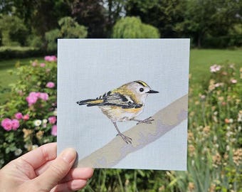 May include: A square card featuring a detailed illustration of a goldcrest bird perched on a branch. The bird has a yellow crown stripe, black eye markings, and olive-green wings with white bars, set against a solid light blue background.