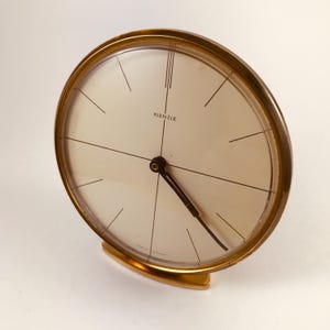 May include: A vintage gold-coloured Kienzle clock with a cream face and black hands. The clock has a circular shape and a curved base. The clock face features black lines indicating the hours and the brand name "KIENZLE".