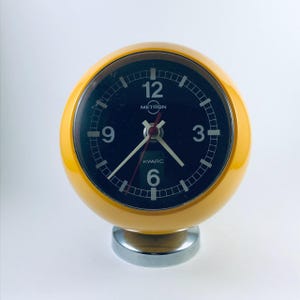 Puede incluir: Un reloj despertador Metron Kwarc vintage con una caja esférica de color amarillo brillante y una esfera negra. El reloj tiene manecillas y números blancos y una base cromada. La esfera del reloj muestra las marcas Metron y Kwarc.