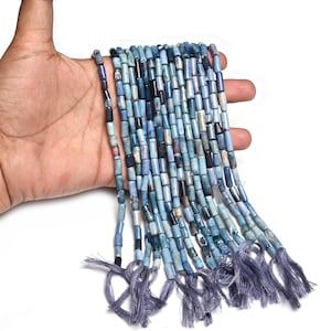 May include: Strands of blue cylindrical beads with varying shades of blue and gray. Each strand is finished with a tassel of blue thread. The beads are held in a hand against a white background.