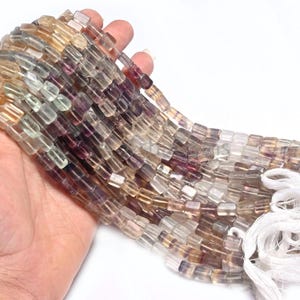 Puede incluir: Una hebra de cuentas de fluorita multicolores, con tonos de morado, verde y amarillo. Las cuentas son de forma rectangular y tienen un acabado liso y pulido. Las cuentas están enhebradas en un hilo blanco y están listas para ser utilizadas para la elaboración de joyas.