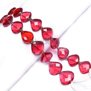 Puede incluir: Dos hilos de cuentas de piedras preciosas rojas translúcidas facetadas. Las cuentas son de corte cuadrado con bordes redondeados y están ensartadas con pequeñas cuentas espaciadoras de color plateado. El fondo es blanco.