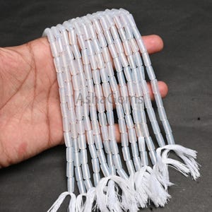 Puede incluir: Múltiples hebras de cuentas cilíndricas translúcidas blanquecinas, cada una con una borla blanca. Las cuentas son de forma y tamaño uniformes. El texto "AshaGems" es visible en la imagen.