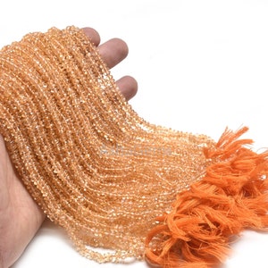 Puede incluir: Un puñado de pequeñas cuentas de color naranja claro, ensartadas, con una borla de color naranja brillante. Las cuentas son de un color claro y translúcido. Las cuentas se sostienen en una mano sobre un fondo blanco.