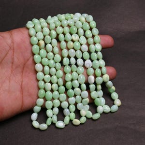 Könnte beinhalten: Eine Kette aus hellgrünen, oval geformten Perlen. Die Perlen haben eine glatte, polierte Oberfläche und variieren leicht in der Farbe, wobei einige weißer erscheinen. Die Perlen sind aufgefädelt und bilden eine Halskette oder ein Armband.