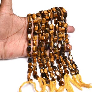 May include: Strands of polished tiger eye gemstone beads in shades of brown and gold. The beads are rectangular and strung together with yellow thread. The gemstones have a natural, striped appearance.