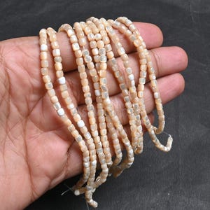 Puede incluir: Hebras de pequeñas cuentas cuadradas de color melocotón con detalles blancos. Las cuentas están ensartadas, formando múltiples líneas. Las cuentas se sostienen en una mano sobre un fondo oscuro. Ideal para la fabricación de joyas.