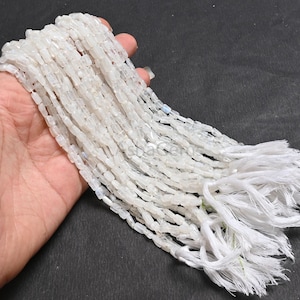 Peut inclure: Un fil de perles blanches rectangulaires avec un pompon. Les perles sont tenues dans une main sur un fond sombre. Les perles sont enfilées ensemble et le pompon est fait de fils blancs.