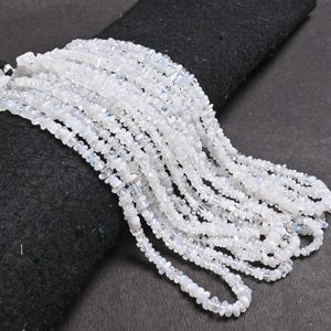 Puede incluir: Una hebra de cuentas de piedra lunar blanca, con una forma lisa y ligeramente irregular. Las cuentas están enhebradas en un hilo transparente y la hebra está sobre un fondo de tela negra.