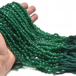 May include: A strand of polished, oval-shaped green beads. The beads are strung together, with a tassel of green thread at the end. The beads are held in a hand against a white background. The text "AshaGems" is visible.