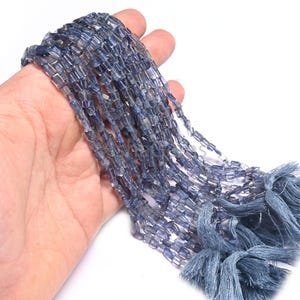 Puede incluir: Una hebra de cuentas de piedra preciosa de cianita azul, cada cuenta tiene una forma rectangular con un borde ligeramente redondeado. Las cuentas están enhebradas en un cordón delgado y tienen un borla al final.