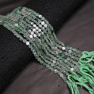 May include: Strands of light green gemstone beads with a mix of oval and round shapes. The beads are strung together, with green tassels at the ends. The beads are displayed on a black surface.