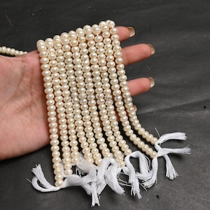 Puede incluir: Múltiples hilos de cuentas de perlas de color crema. Cada hilo está terminado con una borla blanca. Las perlas de forma ovalada tienen una superficie lisa y brillante. Las cuentas se muestran sobre un fondo oscuro. Las perlas miden aproximadamente 0,5 cm de diámetro.