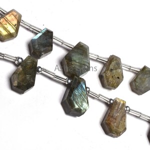 May include: A strand of faceted labradorite gemstone beads in a shield shape, featuring iridescent flashes of blue, gold, and green. The stones are strung on a silver-toned wire with small metallic spacer beads between each piece, displaying the text Asha Gems.