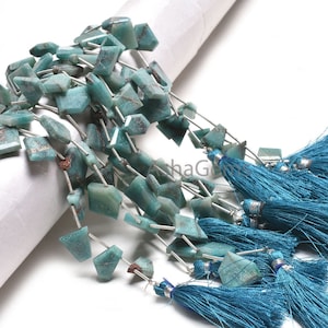 Peut inclure: Des rangées de perles de pierres précieuses turquoise aux formes irrégulières et facettées. Les perles sont enfilées sur du fil argenté et agrémentées de pompons bleu sarcelle. Les pierres ont un aspect moucheté.