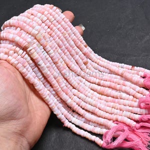 Könnte beinhalten: Stränge aus rosa Opal-Edelsteinperlen. Die Perlen sind klein, flach und scheibenförmig, mit einem zarten rosa Farbton und subtilen Farbvariationen. Die Stränge werden mit rosa Faden zusammengehalten, und die Enden sind mit einer Quaste versehen.
