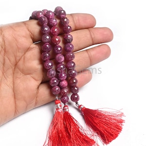 Op de afbeelding: Strengen gefacetteerde, dieprode edelsteen kralen met bijpassende rode kwastjes. De kralen zijn rond en hebben een glanzende, gepolijste afwerking. De kwastjes zijn gemaakt van fijne, zijdeachtige draden en zijn bevestigd met zilverkleurige metalen accenten.