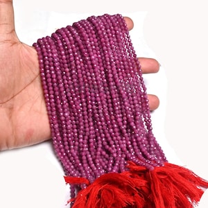 May include: A long strand of small, faceted, deep magenta-colored gemstone beads arranged in multiple parallel rows. The beads have a uniform, round shape and a reflective surface. The strand is finished with a bright red tassel at the bottom end.