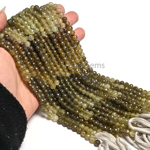 Puede incluir: Una hebra de cuentas de piedras preciosas facetadas de color verde y marrón. Las cuentas son redondas y tienen una forma ligeramente irregular. La hebra mide aproximadamente 40 cm de largo y está enhebrada en un cordón blanco.