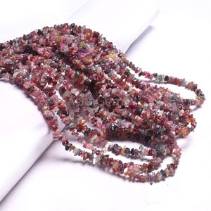 Peut inclure: Plusieurs brins de perles en éclats de tourmaline multicolores. Les perles présentent un mélange de teintes rouges, roses, vertes et brunes. Les éclats sont enfilés ensemble, créant un aspect texturé et naturel. Les perles sont présentées sur une surface blanche.