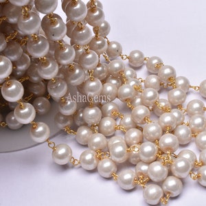 May include: A strand of white faux pearl beads strung on a gold chain. The beads are round and smooth, and the chain is delicate and shiny.