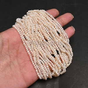 Puede incluir: Una colección de delicadas hebras de perlas blanquecinas. Las perlas son pequeñas y redondas, creando un efecto texturizado y en capas. Las hebras se sostienen en una mano, mostrando el brillo y el color natural de las perlas.