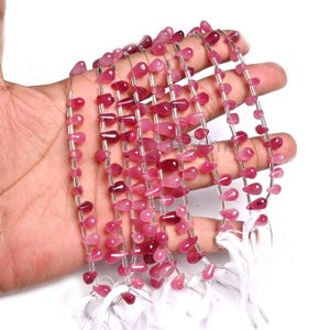 Pode incluir: Múltiplos fios de contas em forma de gota em vários tons de rosa e magenta. As contas são translúcidas com um acabamento liso e polido, enfiadas num fio branco fino, criando uma coleção de delicados componentes para joalharia.